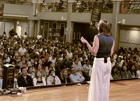 Reyna Ana en el escenario, de espaldas, ante una sala llena de asistentes.
